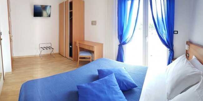Camera da letto con letto matrimoniale blu, finestre con tende azzurre, scrivania e TV.