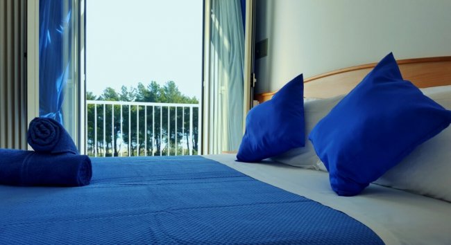 Camera con letto matrimoniale, cuscini e coperte blu, vista su balcone con alberi.