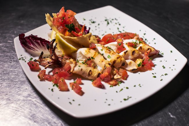Calamari grigliati con pomodori e prezzemolo su un piatto bianco.