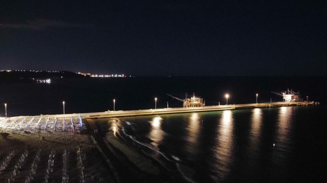 Molo illuminato di notte con riflessi sull'acqua, vista dall'alto.