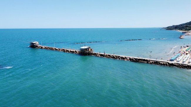 Lungo molo di legno che si estende nel mare, affiancato da una spiaggia con ombrelloni.