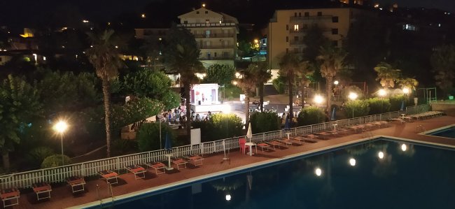 Piscina notturna illuminata con palme e edifici vicini.