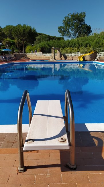 Pedana per tuffi di una piscina all'aperto, con area circostante verde e alberata.
