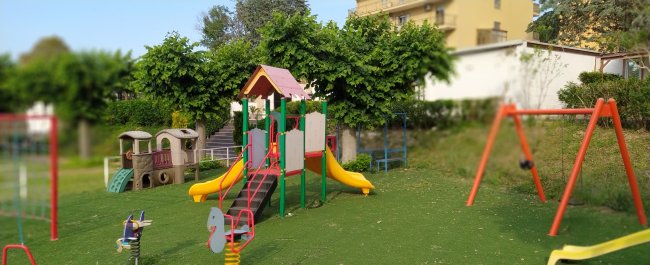 Parco giochi con scivolo giallo, altalene e casetta su prato verde.