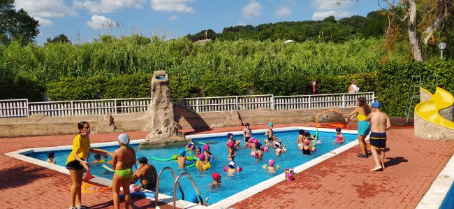 Bambini con cuffie colorate nuotano in una piccola piscina, circondati da istruttori.