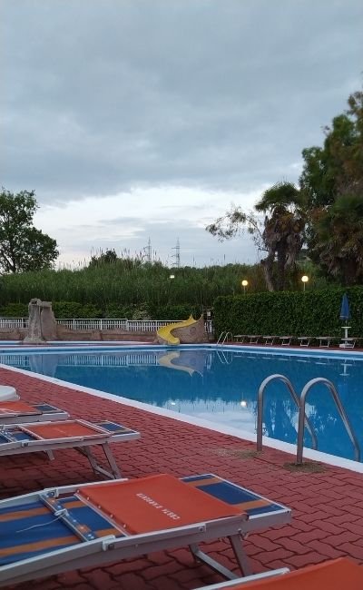 Piscina con sdraio arancioni e scivolo giallo, circondata da alberi e cielo nuvoloso.