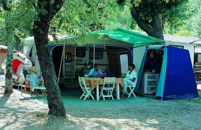 Camper all'ombra con tavolo e sedie all'aperto.