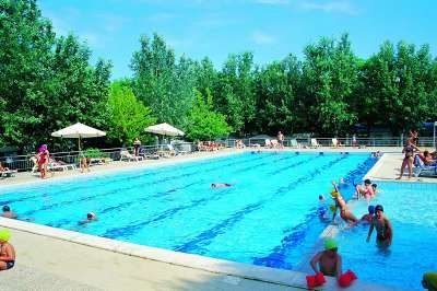 Una piscina con diverse persone che nuotano e giocano, circondata da alberi e lettini.