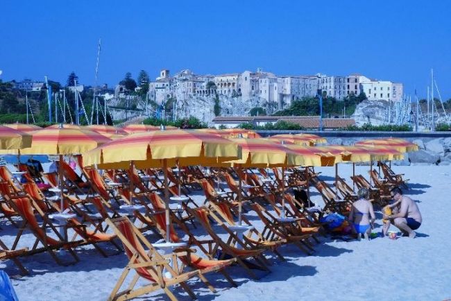 Spiaggia sabbiosa con file di ombrelloni gialli e sdraio. Sullo sfondo, una città sulla scogliera.