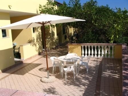 Terrazza con ombrellone e sedie bianche, circondata da muretti e vegetazione.