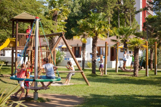 Parco giochi per bambini con altalene e palme.