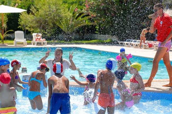 Bambini giocano in piscina, schizzando acqua, sotto la supervisione di un adulto in costume rosso.