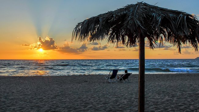 Spiaggia al tramonto con due sedie a sdraio sotto un ombrellone di paglia.