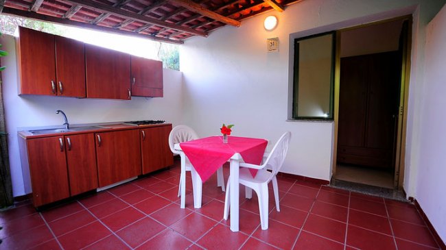 Cucina esterna con tavolo rosso, sedie bianche e credenze in legno.