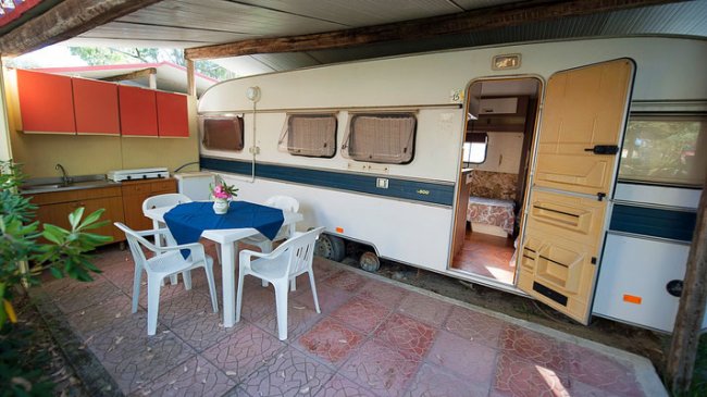 Una roulotte con una veranda arredata con tavolo, sedie bianche e un piccolo angolo cottura.