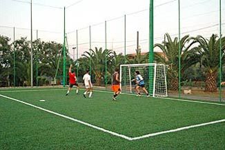 Una partita di calcetto in corso su un campo con erba sintetica e palme sullo sfondo.
