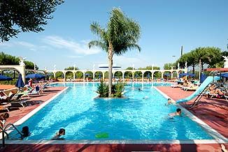 Piscina all'aperto con una palma centrale e scivolo, circondata da lettini e persone che nuotano.