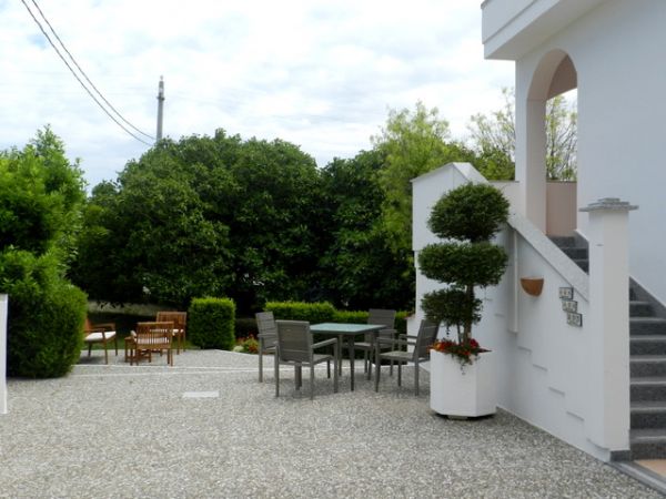 Cortile con tavolo e sedie, circondato da alberi e siepi.