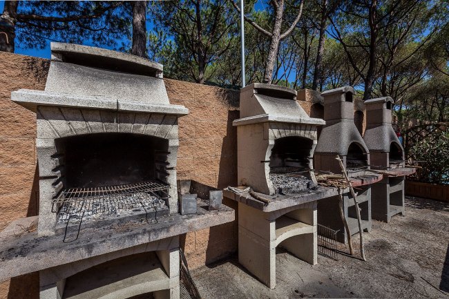 Un'area barbecue con cinque griglie in muratura sotto un cielo blu e alberi.