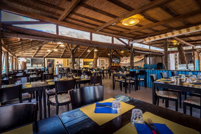 Un ristorante all'aperto con tetto di paglia, tavoli apparecchiati con tovagliette gialle e posate.