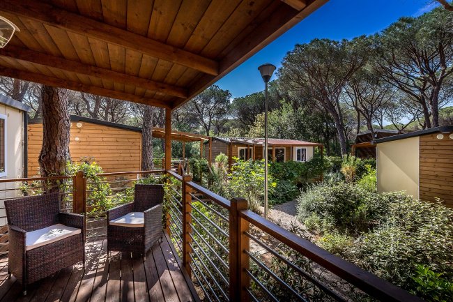 Veranda in legno con sedie, circondata da vegetazione e bungalow in legno.