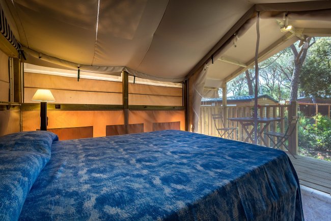 Interno di una tenda con letto matrimoniale, copriletto blu e lampada accesa.