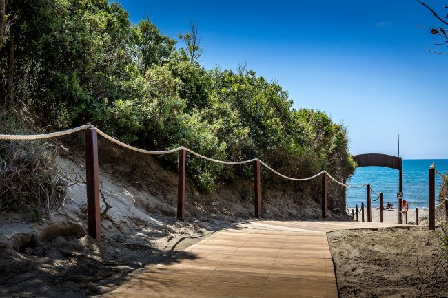 Sentiero in legno tra dune sabbiose conduce al mare, circondato da vegetazione.
