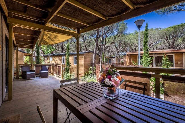 Veranda in legno con tavolo e fiori, circondata da bungalow tra gli alberi.