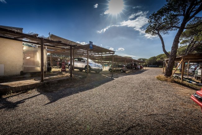 Parcheggio ghiaioso ombreggiato con auto sotto tettoie.