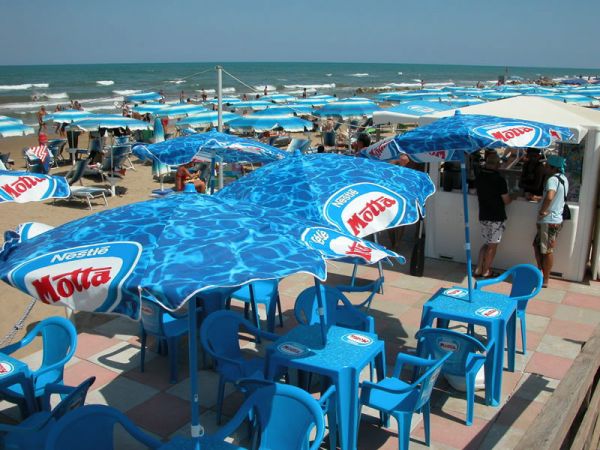 Ombrelloni e sedie blu su una terrazza sul mare con la spiaggia affollata sullo sfondo.