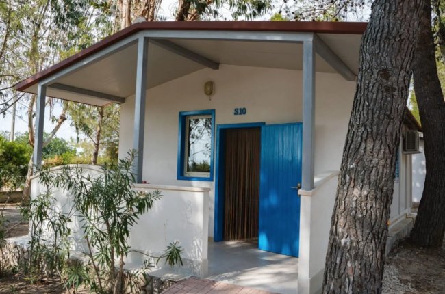 Bungalow bianco con porta blu, circondato da alberi e siepi.