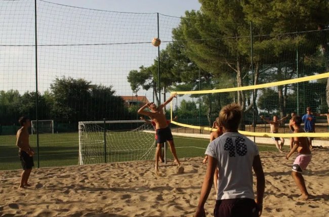 Persone giocano a beach volley su un campo sabbioso con reti e alberi intorno.