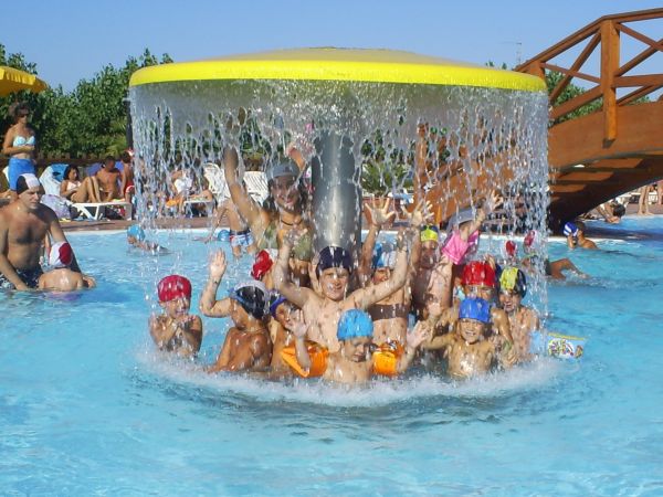 Bambini sorridenti con cappelli colorati giocano sotto una cascata in piscina.