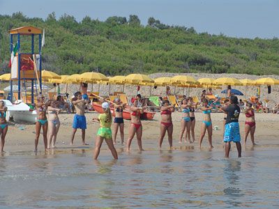 Persone in costume da bagno fanno aerobica in acqua vicino a ombrelloni gialli sulla spiaggia.