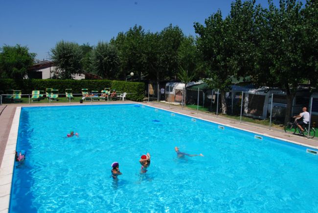 Una piscina con bambini che nuotano, circondata da alberi e sedie a sdraio.