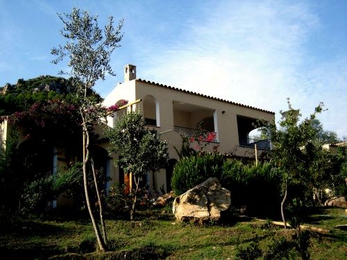 Casa mediterranea con balconi, circondata da alberi e arbusti.