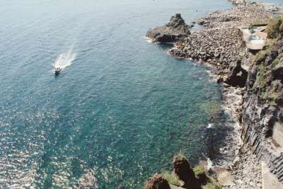 Una costa rocciosa con mare blu e una barca che lascia una scia sull'acqua.