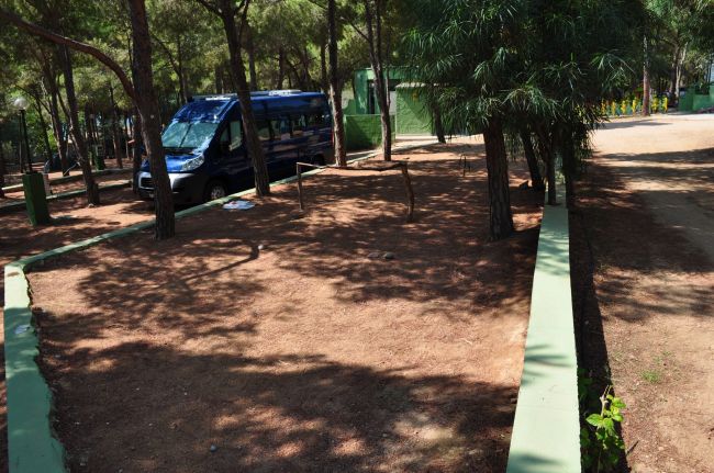Area campeggio ombreggiata con terreno di terra battuta e un furgone blu tra gli alberi.