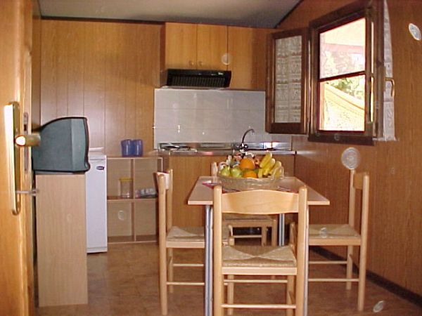 Cucina con tavolo rettangolare, quattro sedie, frigorifero piccolo, e mensole. Finestra aperta.