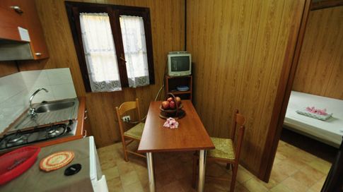 Cucina con tavolo marrone, due sedie, TV su scaffale, e lavello. Camera da letto visibile.