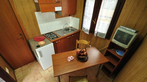 Piccola cucina con tavolo di legno, sedie, mini frigo, lavandino, fornelli e TV su mensola.