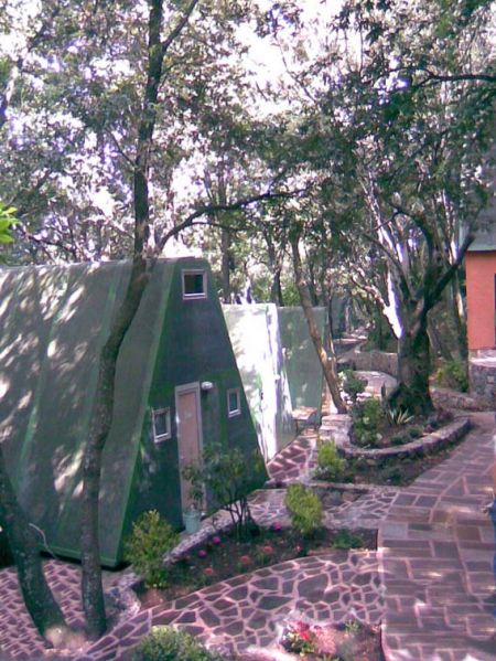 Case verdi a forma triangolare allineate lungo un sentiero pavimentato, circondate da alberi.