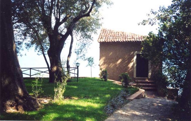Una piccola casa di pietra con tetto di tegole, circondata da alberi e prato verde.