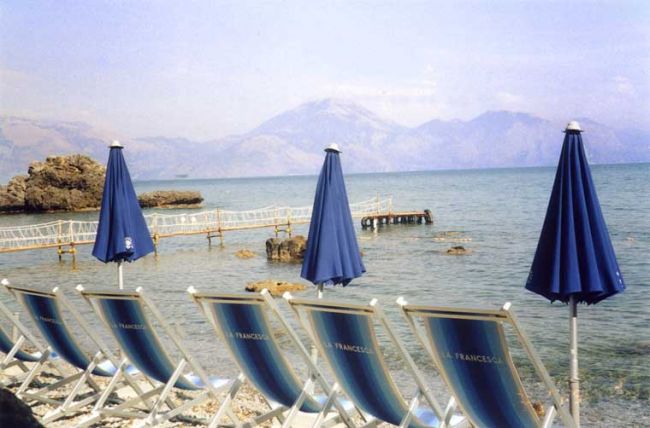 Spiaggia con sedie a sdraio blu e ombrelloni, vista sul mare e montagne sullo sfondo.