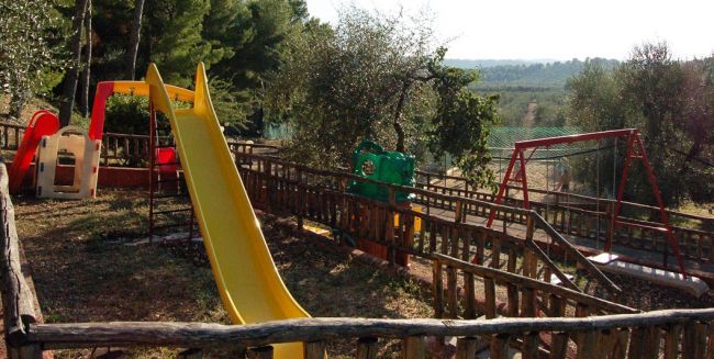 Un parco giochi recintato con uno scivolo giallo e altalene tra gli alberi.