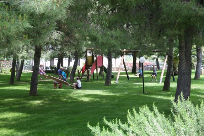 Bambini giocano in un parco verde con altalene e alberi.