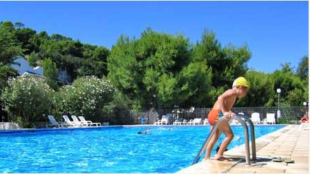 Un bambino con cuffia gialla esce da una piscina all'aperto circondata da alberi.