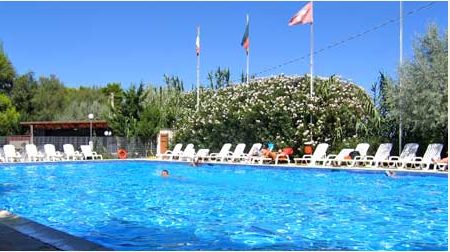Piscina all'aperto con sedie a sdraio, bandiere e alberi sullo sfondo.