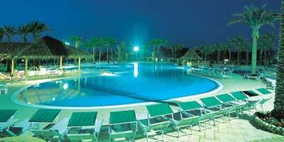 Grande piscina notturna con lettini, circondata da palme e capanne di paglia.