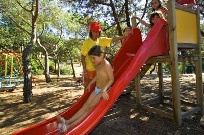 Bambini che giocano su uno scivolo rosso in un parco giochi ombreggiato da alberi.